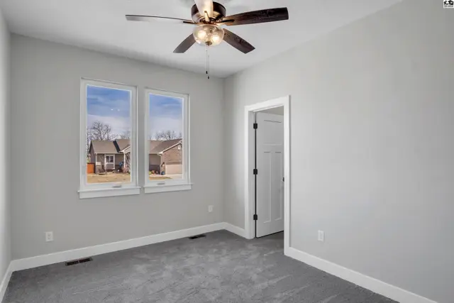 Bright bedroom with gray carpet, white trim, and a ceiling fan, overlooking a suburban street.