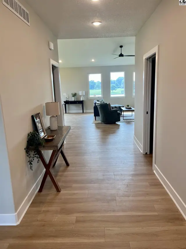 Hallway with wood-look flooring leads to a bright living area with large windows and a ceiling fan.