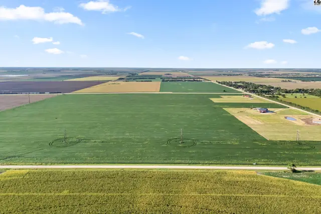Expansive agricultural land with green fields, yellow crops, and a distant farmhouse under a blue sky.