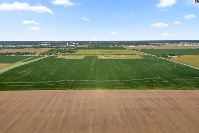 Expansive agricultural fields with green crops and brown soil stretch towards a distant town under a blue sky.