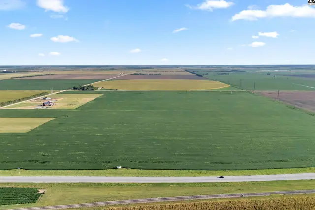 Expansive agricultural land with green fields, farm buildings, and a highway under a clear blue sky.