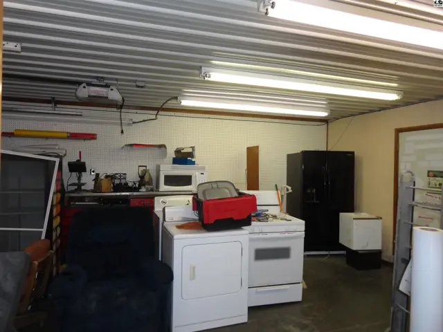 Spacious garage with appliances, storage, and fluorescent lighting under a corrugated metal ceiling.