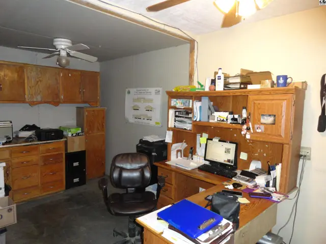 A cluttered home office with a large wooden desk, built-in cabinets, and a ceiling fan.