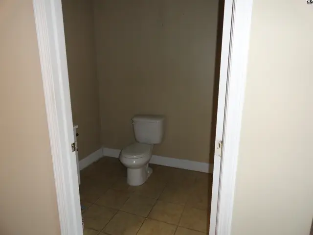 A simple toilet sits in a small, tiled room with beige walls, accessed through a doorway.