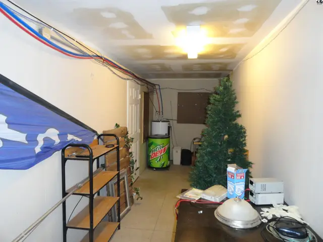 Hallway with unfinished ceiling, exposed wires, shelving, a Christmas tree, and a mini-fridge.