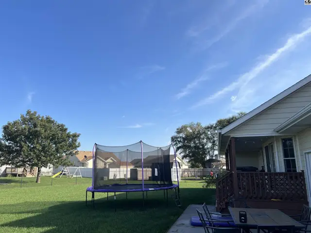 A fenced backyard features a trampoline, a wooden deck, and a patio with outdoor seating.