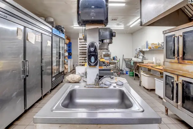 Commercial kitchen with stainless steel sink, mixers, ovens, and large refrigerators.