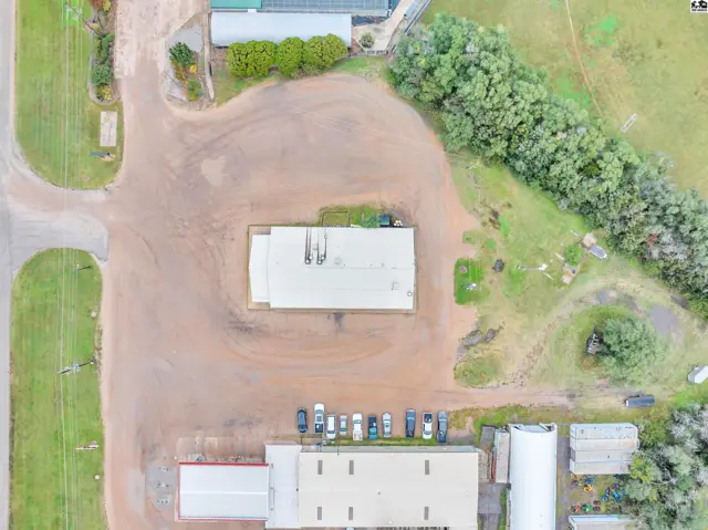 Aerial view of industrial buildings with gravel lot, parking, and surrounding greenery.