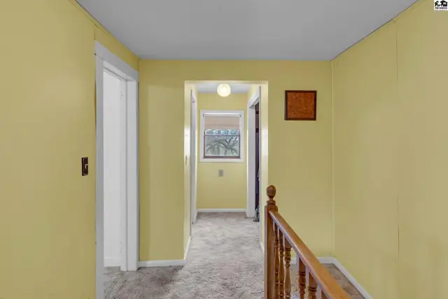 Hallway with yellow walls, carpeted floor, and wooden banister leading to a window.