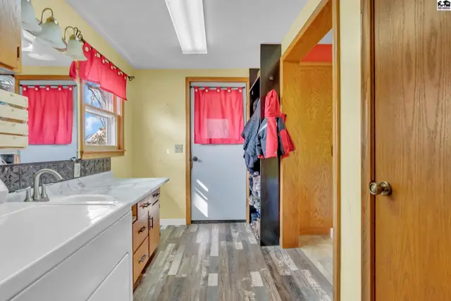 Kitchen with double sink, light wood cabinets, and gray wood-look flooring, leading to a hallway with a coat rack.