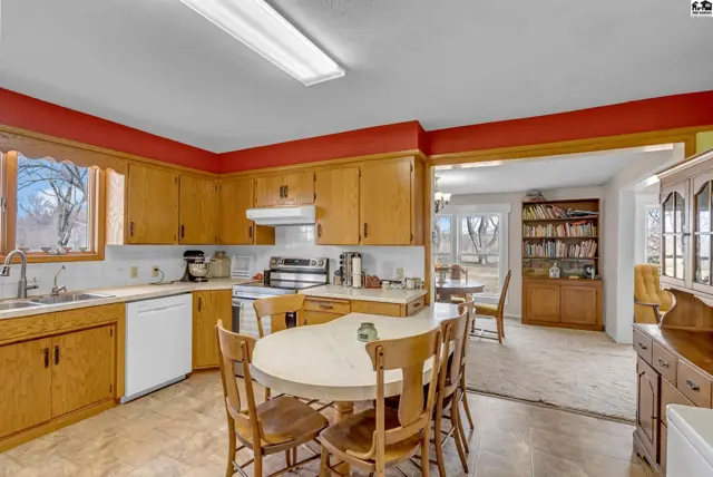 Spacious kitchen with light wood cabinets, white appliances, and a central dining table, opening to a sunlit dining area.