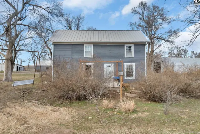 Gray clapboard farmhouse with a metal roof, surrounded by bare trees and overgrown bushes.