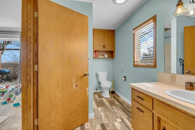 Light blue bathroom with wood vanity, laminate flooring, and a wooden door leading to another room.