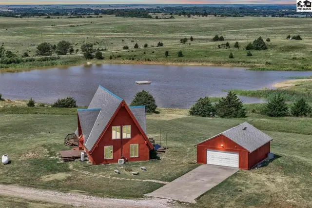 Red A-frame house and detached garage sit on a grassy lot overlooking a large pond.