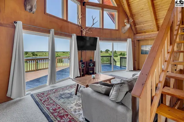Spacious living room with vaulted wood ceilings, large windows, and a rustic staircase overlooking a scenic landscape.