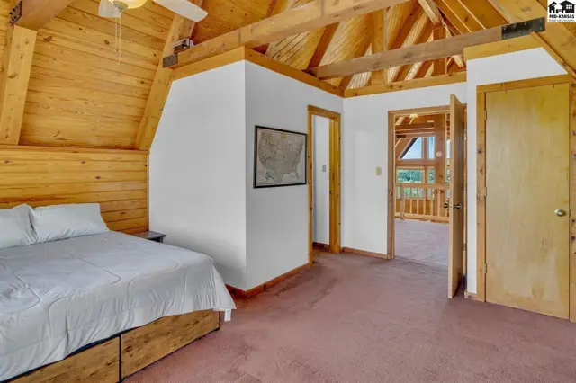 Spacious bedroom with vaulted wood-beamed ceiling, white walls, and plush carpet, opening to a balcony view.