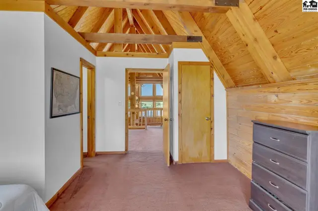 Rustic attic bedroom with exposed wood beams, paneled walls, and a view into a sunlit hallway.