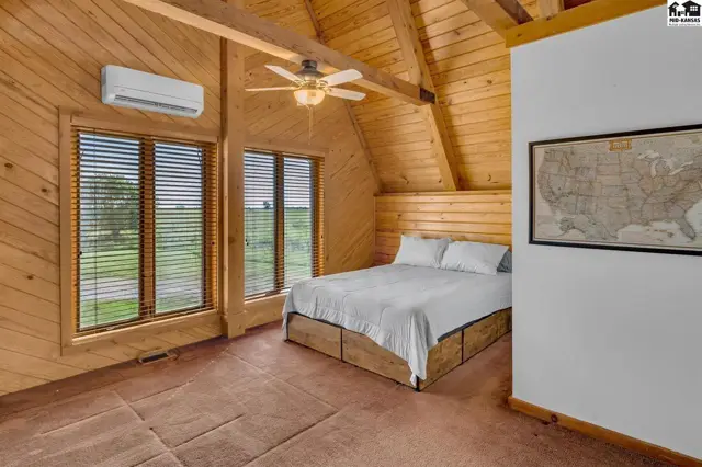 Cozy bedroom with wood-paneled walls, vaulted ceiling, and large windows overlooking a rural landscape.