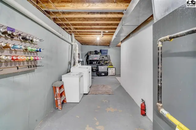 Unfinished basement with laundry machines, storage shelves, and exposed wooden ceiling joists.