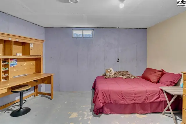Unfinished basement bedroom with a wooden desk, stool, and a bed with burgundy linens.