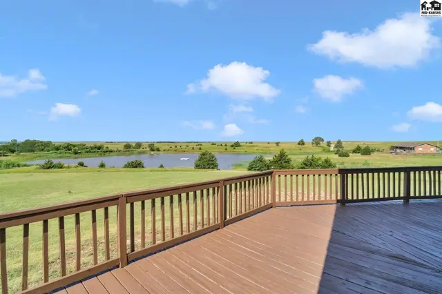 Spacious wooden deck with brown railings overlooks a serene lake and rolling green landscape under a blue sky.