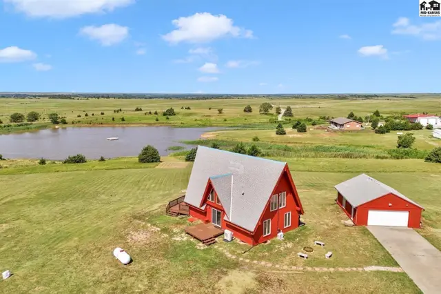 Red A-frame house with a detached garage sits on a grassy lot beside a tranquil pond.