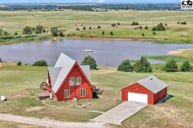 Red A-frame house and detached garage sit on a grassy lot overlooking a large pond.