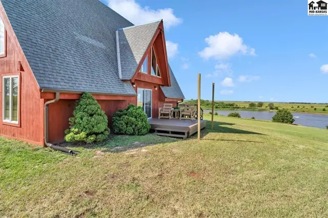 Red A-frame home with a wooden deck overlooks a scenic lake under a bright blue sky.