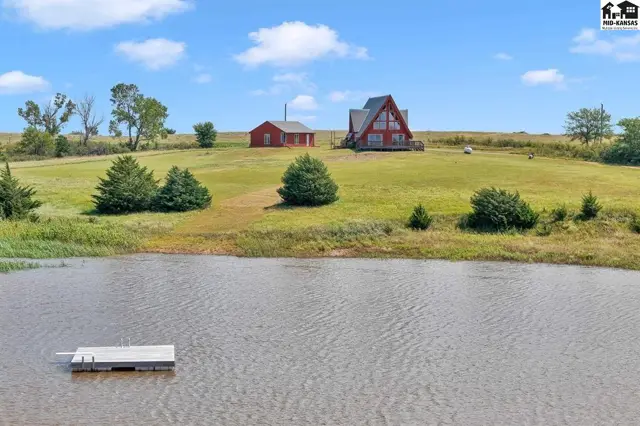 A-frame house and red barn sit on a grassy hill overlooking a calm pond with a small dock.