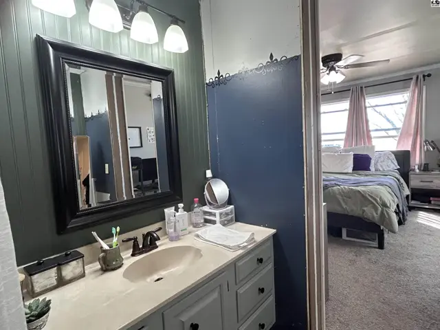 Bathroom vanity with a dark mirror and beadboard walls, opening to a bedroom with a bed and ceiling fan.