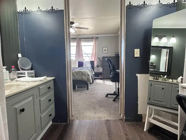 View into a bedroom from a bathroom with gray vanities, dark blue walls, and a ceiling fan.