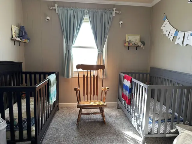 Nursery with two cribs, a wooden rocking chair, and light blue curtains.