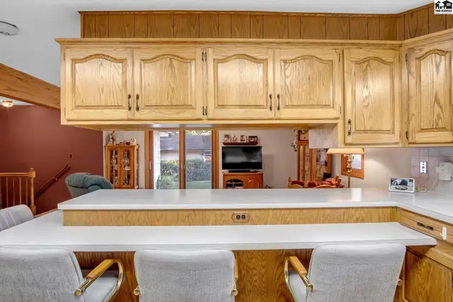 Kitchen features light wood cabinetry, white countertops, and a breakfast bar with upholstered stools.