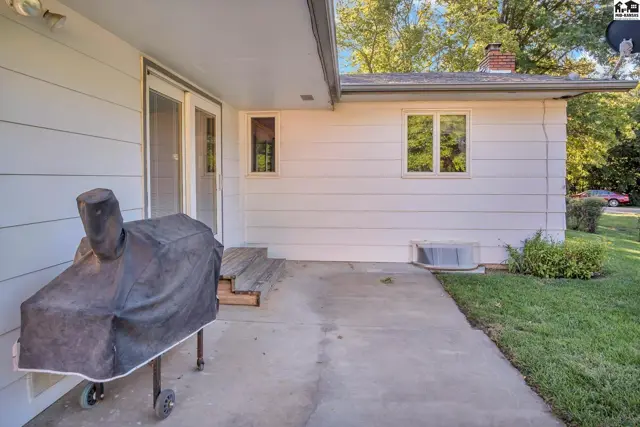 Concrete patio with a covered grill sits beside a white-sided home with a brick chimney.