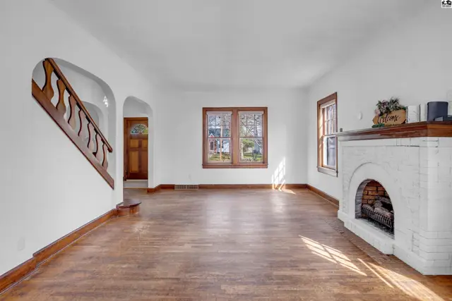 Spacious living room with white brick fireplace, hardwood floors, and wooden staircase.
