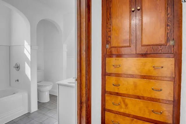 A bathroom with white tub and toilet is visible through a doorway next to a tall wooden cabinet with drawers.