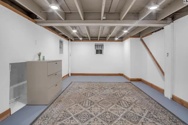 Unfinished basement with exposed joists, white walls, a large rug, and a dresser.