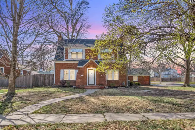 Charming brick two-story home with white shutters, a white door, and mature trees in a suburban setting.