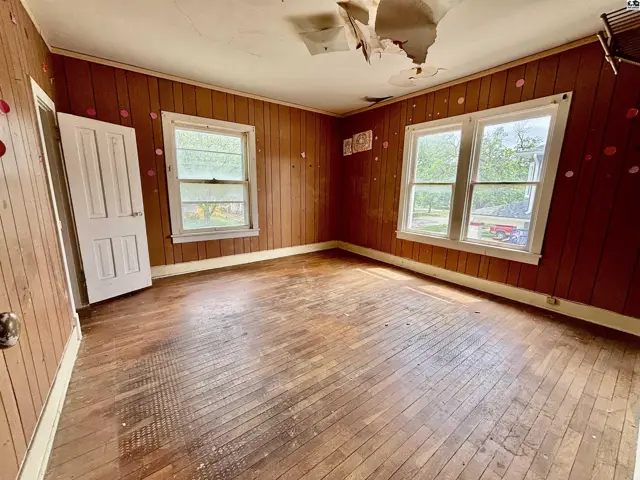 Empty room with wood-paneled walls, hardwood floors, and two windows, showing signs of needed renovation.