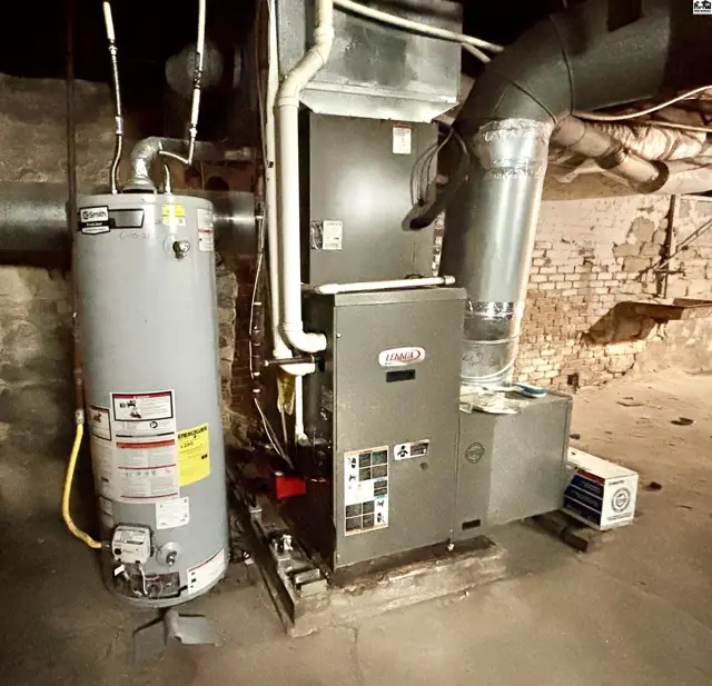 Furnace and water heater with exposed ductwork and brick wall in a basement.