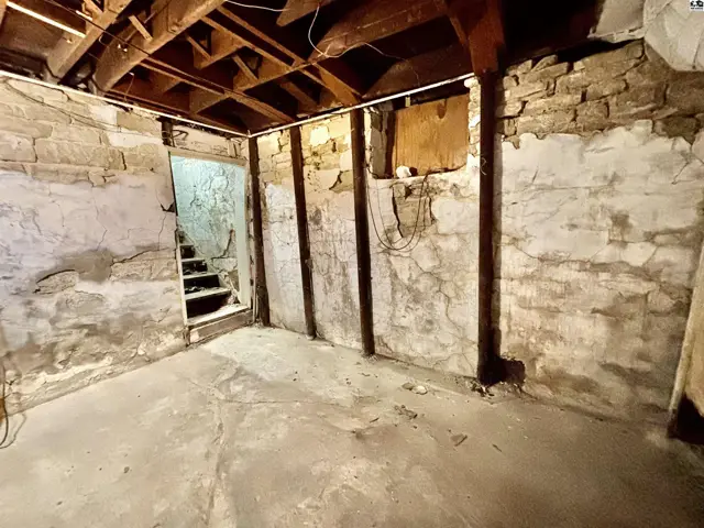Rustic basement with exposed stone walls, wooden beams, and a concrete floor leading to stairs.