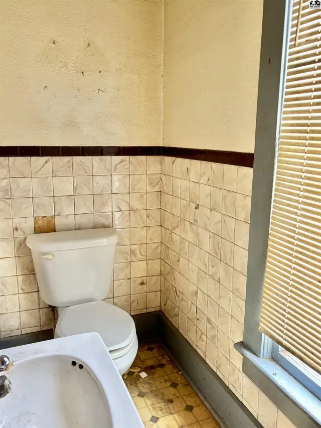 A bathroom with a white toilet, sink, and tiled walls, featuring a patterned floor and a window with blinds.