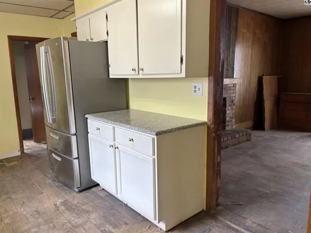 Kitchen area with stainless steel refrigerator, white cabinets, granite countertops, and wood-paneled walls.