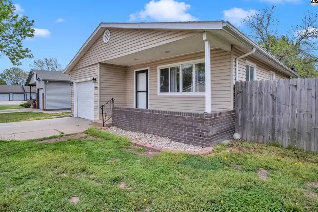 Tan vinyl-sided house with brick foundation, covered porch, and attached garage.