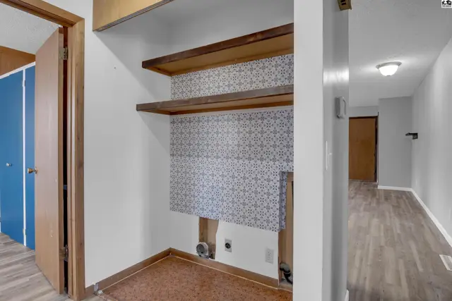 Laundry nook with patterned wallpaper, wooden shelves, and a view of a hallway with wood-look flooring.