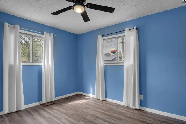 Bright bedroom with blue walls, wood-look flooring, white curtains, and a ceiling fan.