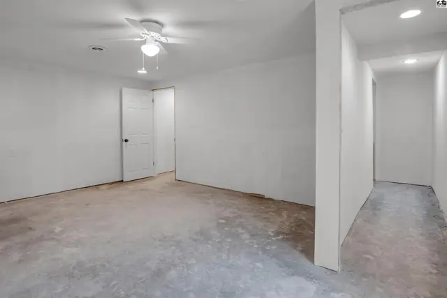 Unfinished basement room with concrete floors, white walls, a ceiling fan, and a hallway leading to more space.