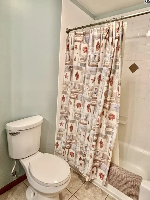 A bathroom with a white toilet, tiled floor, and a seashell-patterned shower curtain.