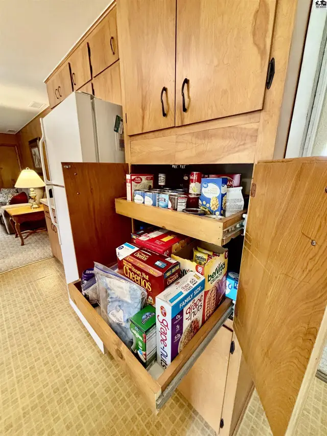 Pantry with pull-out shelves stocked with cereals and canned goods, adjacent to a refrigerator and wood cabinetry.