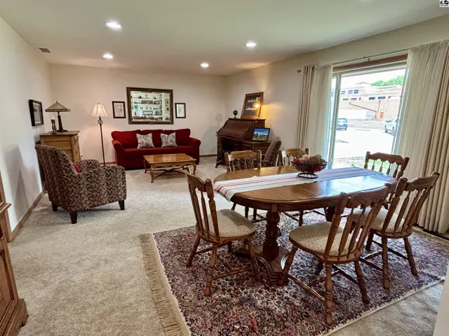 Spacious living and dining area with plush carpet, wood furniture, and a large window.
